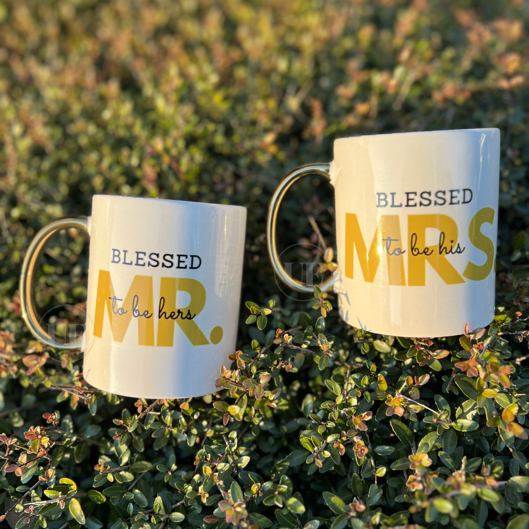 A set (2) 11oz white ceramic mugs with gold handles that read MR. Blessed to be hers and the other says MRS. Blessed to be his.  Mr and Mrs on each mug is a gold color.